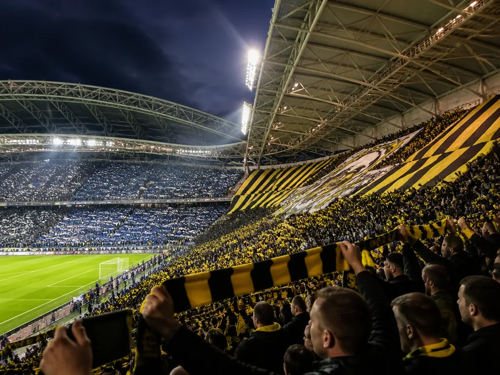 Fullsatt fotbollsstadion med svart-gula och blå-vita supportrar under kraftfull floodljusbelysning i ett Stockholmsderby