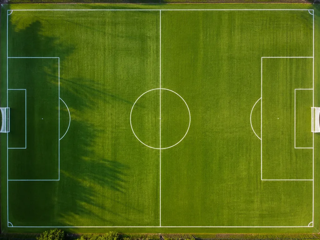 Fotbollsplan sedd uppifrån med tydliga vita linjemarkeringar, straffområde och mittcirkel på grönt gräs